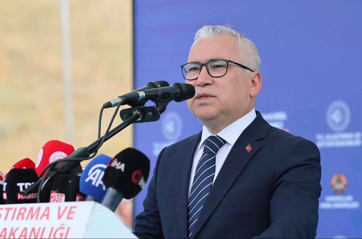 Kürsüde konuşan bir kişi, mikrofonun önünde duruyor ve arka planda mavi bir pankart var.