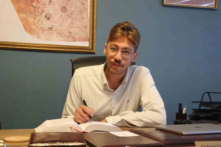 Gözlüklü bir kişi, masada açık bir kitap ve belgelerin yanında kalemle yazı yazıyor.
