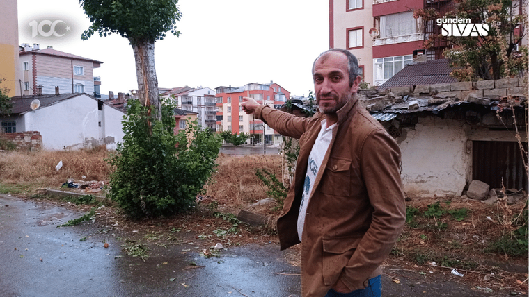 Kahverengi ceketli bir adam, ıslak sokakta elini ilerideki apartmanlara doğru uzatıyor.