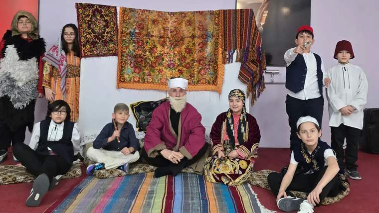 Farklı kıyafetler giymiş çocuklar ve bir yetişkin, renkli bir halının önünde oturuyor.