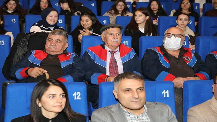 Salonda mavi koltuklarda oturan, kırmızı-mavi cüppeli üç kişi ve arkalarında izleyiciler görülüyor.