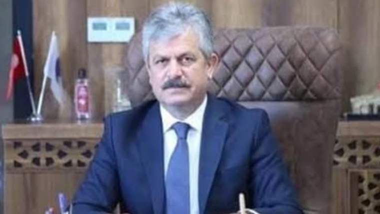 Takım elbiseli, bıyıklı bir kişi ahşap kaplı odada, yüksek sırtlı koltukta masa başında oturuyor.