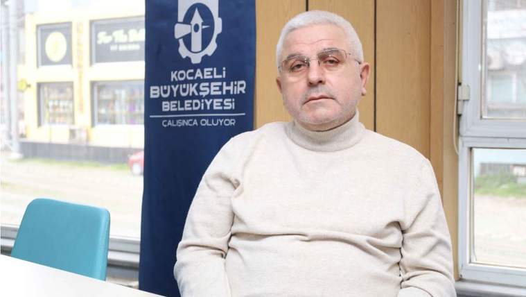 Pencereler önünde oturan beyaz saçlı bir adamın arkasında Kocaeli Büyükşehir Belediyesi yazılı pano bulunuyor.