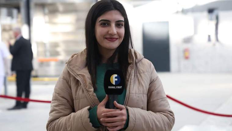 Kahverengi bir mont giymiş, mikrofon tutan genç bir kadın gülümsüyor.