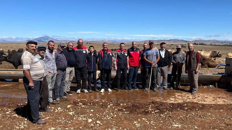 Hayvan içme suyu tesisinin önünde bir grup insan, inekler ve açık bir alan görünmekte.