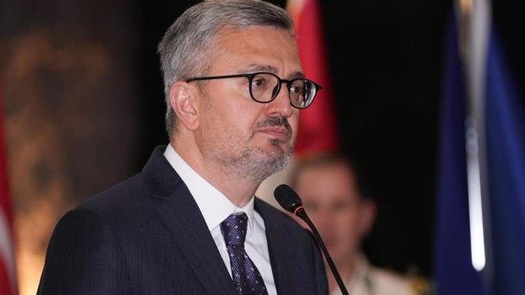 Kürsüde konuşan gözlüklü bir erkek, koyu renk takım elbise ve kravat giyiyor; arkasında bayraklar bulunuyor.