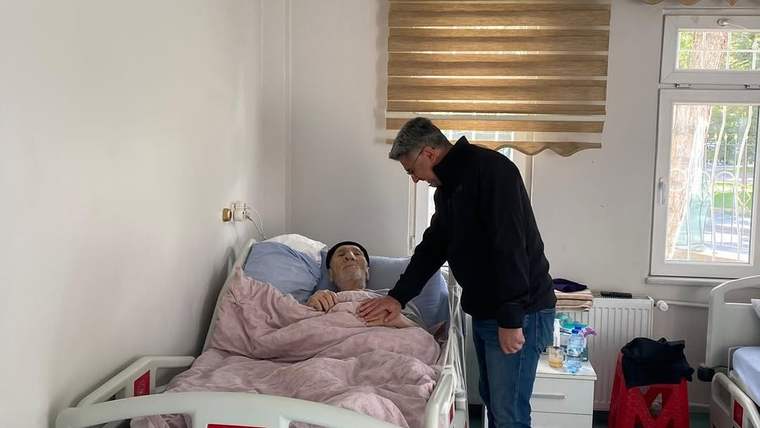 Bakım odasında bir kişi, yatakta uzanan yaşlı bir kişinin elini tutuyor; pencere yanında komodin bulunuyor.