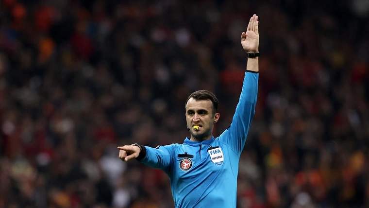 Mavi forması üzerinde FIFA rozeti bulunan hakem, düdük çalarak bir kolunu kaldırıp diğerini uzatıyor.