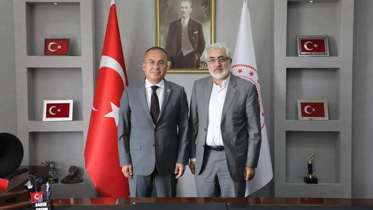 Türk bayrağı ve kurum bayrağı önünde, takım elbiseli iki kişi yan yana duruyor; arkada portre bulunuyor.