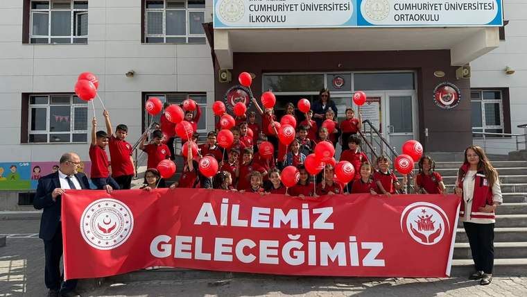 Okul binası önünde kırmızı balonlar taşıyan çocuklar ve yetişkinler, üzerinde “AİLEMİZ GELECEĞİMİZ” yazılı pankart tutuyor.