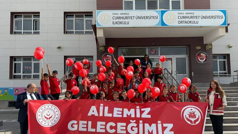 Okul binası önünde kırmızı balonlar taşıyan çocuklar ve yetişkinler, üzerinde “AİLEMİZ GELECEĞİMİZ” yazılı pankart tutuyor.