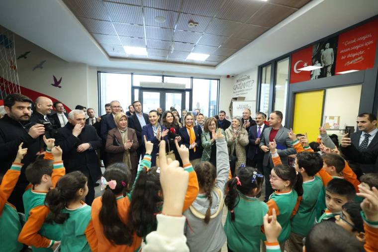 Bir grup çocuk, el kaldırarak bir etkinlikte yer alıyor; arka planda yetişkinler ve bir kapı görünmekte.