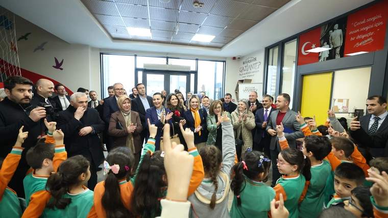 Bir grup çocuk, el kaldırarak bir etkinlikte yer alıyor; arka planda yetişkinler ve bir kapı görünmekte.