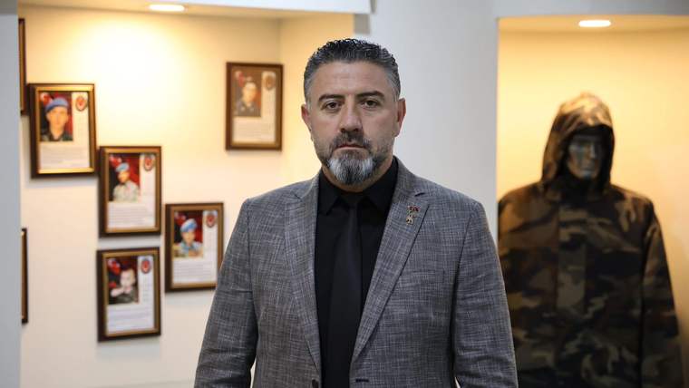 Siyah kravat takan, gri ceket giymiş bir adam, arkasında çerçevelenmiş fotoğraflar ve bir kıyafet sergileniyor.