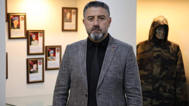 Siyah kravat takan, gri ceket giymiş bir adam, arkasında çerçevelenmiş fotoğraflar ve bir kıyafet sergileniyor.