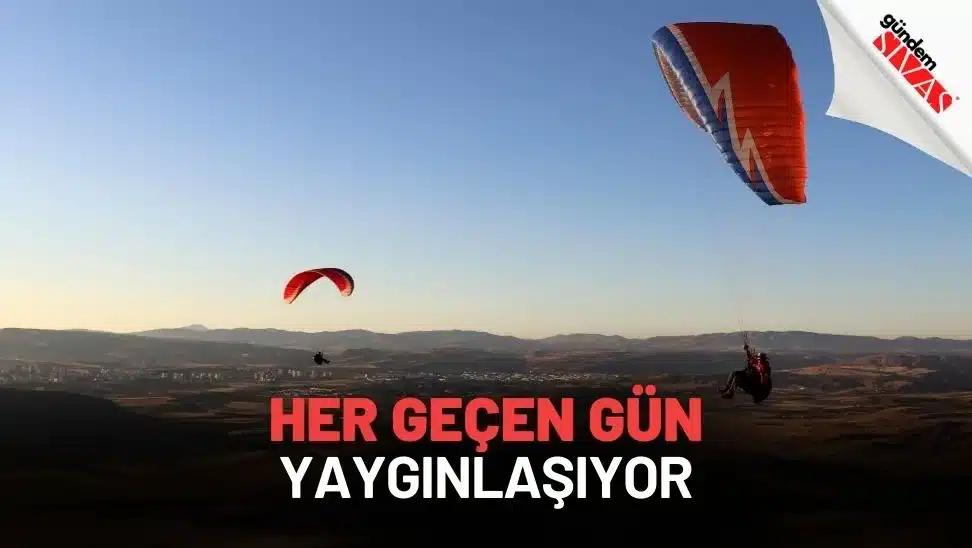 Gökyüzünde paraşütle uçan iki kişi, arka planda dağlar ve açık bir alan görünmekte.