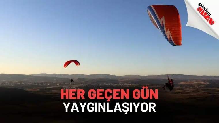 Gökyüzünde paraşütle uçan iki kişi, arka planda dağlar ve açık bir alan görünmekte.