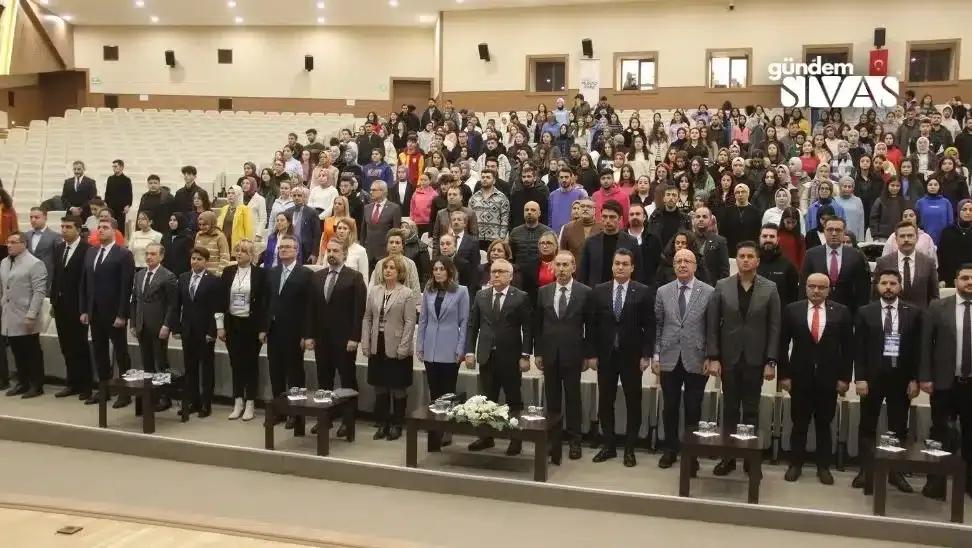 Bir etkinlikte, kalabalık bir grup insan ayakta duruyor, bazıları resmi kıyafetler giymekte.