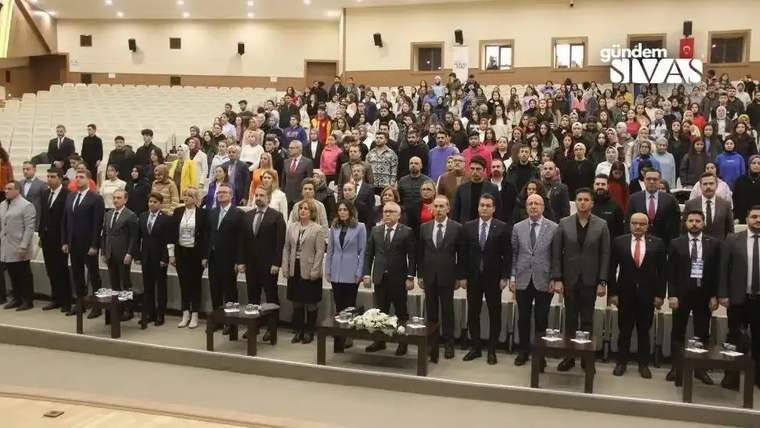 Bir etkinlikte, kalabalık bir grup insan ayakta duruyor, bazıları resmi kıyafetler giymekte.