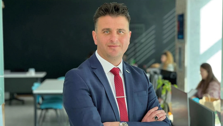 Lacivert takım elbiseli, kırmızı kravatlı bir kişi kollarını bağlayarak iç mekânda kameraya bakıyor.