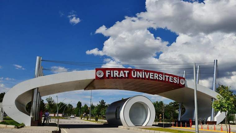 Fırat Üniversitesi yazılı tabelanın altında, beyaz kemerli giriş ve yuvarlak metal yapı bulunan kampüs girişi.