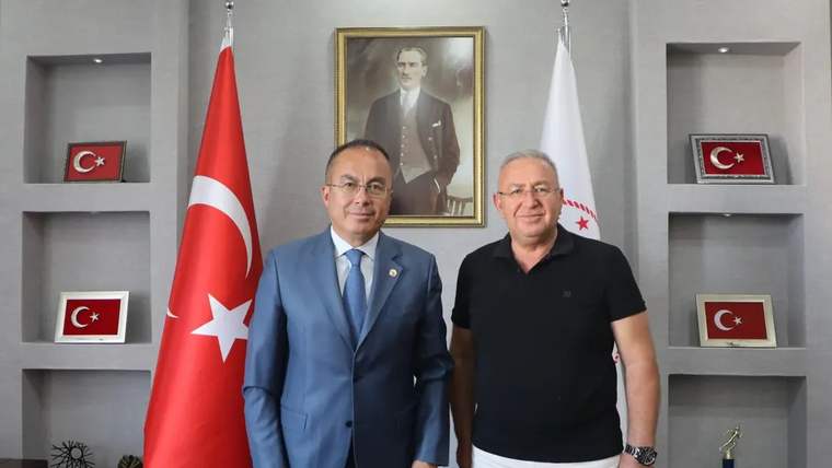 Türk bayrağı ve beyaz flamaların önünde duran iki erkek, duvarda asılı bir portreyle birlikte görülüyor.