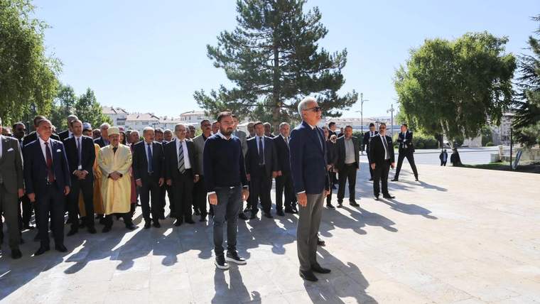 Açık alanda takım elbiseli bir grup insan, sarı giysili bir kişiyle birlikte ağaçların önünde yürüyor.