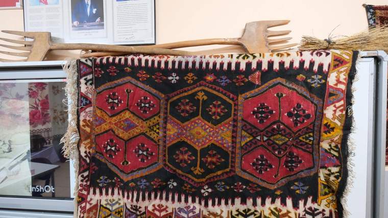 Geometrik desenli renkli bir halı, arkasında çerçeveli belgeler ve üstünde ahşap el aletleriyle sergileniyor.