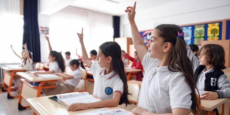Sınıfta elleri havada bekleyen öğrenciler, ders dinliyorlar.