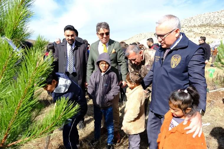 Çocuklar ve yetişkinler, açık alanda çam fidanı çevresinde dururken bir kişi toprağı kazıyor.