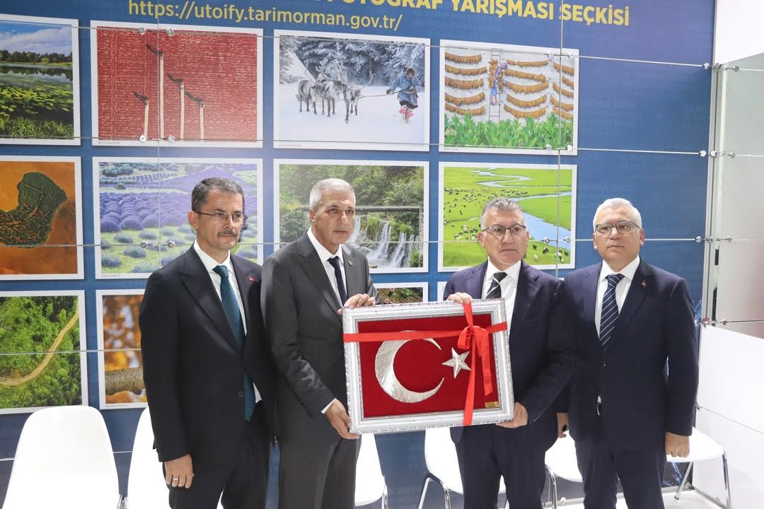 Dört takım elbiseli kişi, üzerinde ay yıldız bulunan kırmızı plaketi fuar standında birlikte tutuyor.
