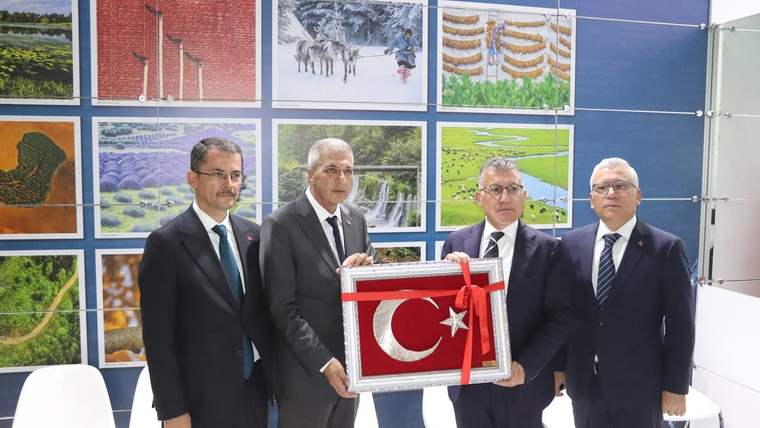 Dört takım elbiseli kişi, üzerinde ay yıldız bulunan kırmızı plaketi fuar standında birlikte tutuyor.