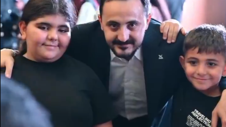 Bir kişi, iki ortaokul öğrencisiyle birlikte gülümsüyor; arka planda kalabalık bir ortam var.