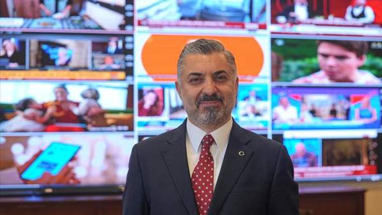 Televizyon ekranlarının önünde duran, koyu renk takım elbiseli ve kırmızı kravatlı bir kişi görülüyor.