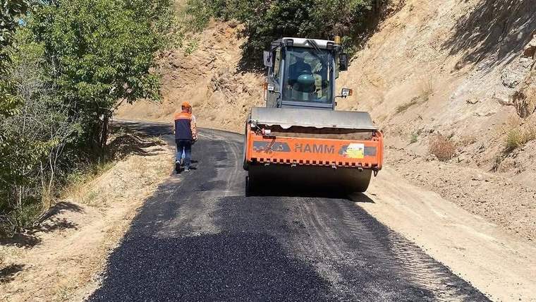 Dağlık bir yolda asfalt silindiri yeni serilmiş koyu renkli yüzeyin üzerinde ilerlerken bir işçi yanında yürüyor.