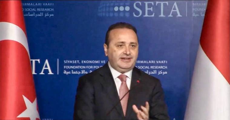 SETA yazılı kürsü önünde takım elbiseli bir kişi konuşurken, iki yanında Türk bayrakları görülüyor.