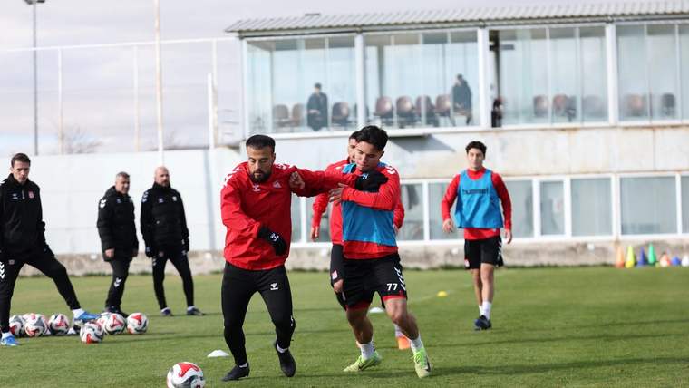 Çim sahada kırmızı formalı iki futbolcu top için mücadele ediyor; arkalarında takım arkadaşları ve camlı bina görülüyor.