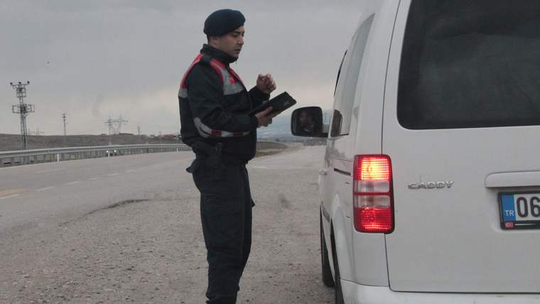 Jandarma üniformalı bir görevli, yol kenarında duran beyaz minibüsün yanında elindeki cihaza bakıyor.