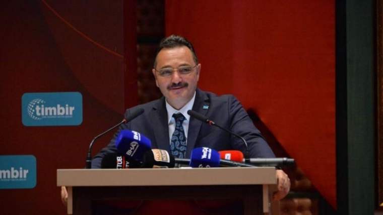 Kürsüde konuşan bıyıklı bir kişi, önündeki mikrofonlar ve arka plandaki timbir yazısıyla görülüyor.