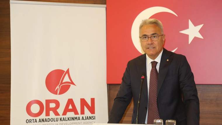 Kürsüde konuşan takım elbiseli bir kişi, yanında ORAN logosu ve arkasında Türk bayrağı bulunan salonda duruyor.