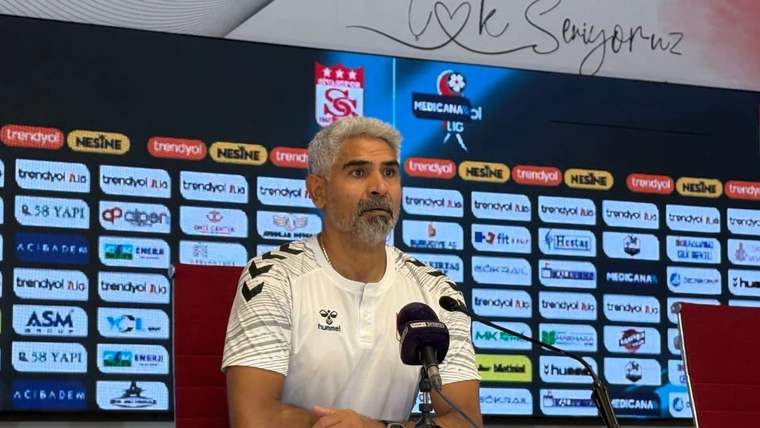 Mikrofonların önünde oturan bir kişi, sponsor logolarının yer aldığı panonun önünde konuşuyor.