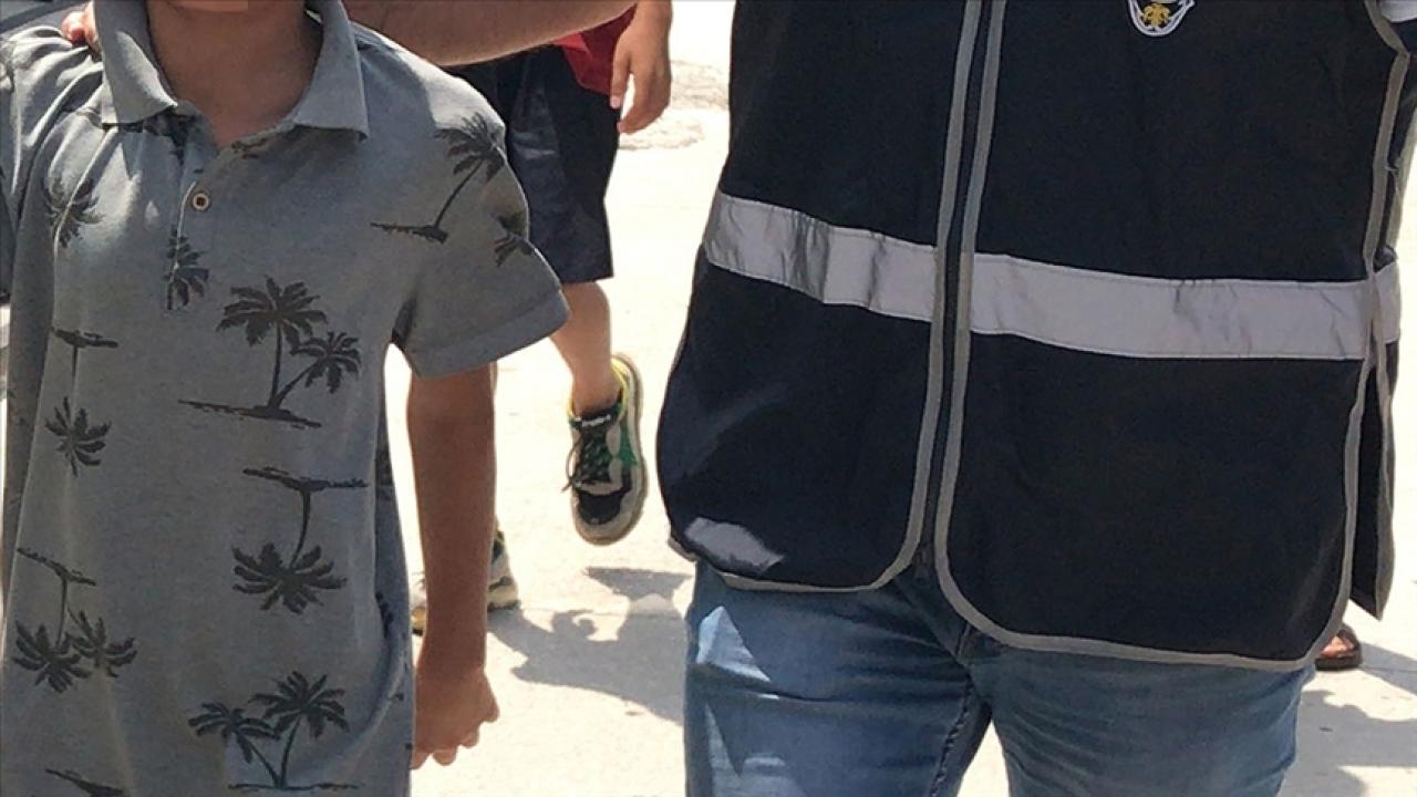 Yüzü görünmeyen bir çocuk, lacivert yelekli bir yetişkinin yanında güneşli bir yolda yürürken görülüyor.