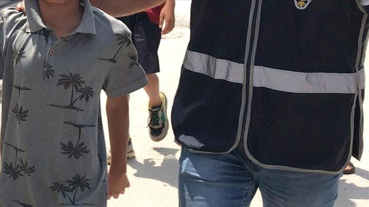 Yüzü görünmeyen bir çocuk, lacivert yelekli bir yetişkinin yanında güneşli bir yolda yürürken görülüyor.