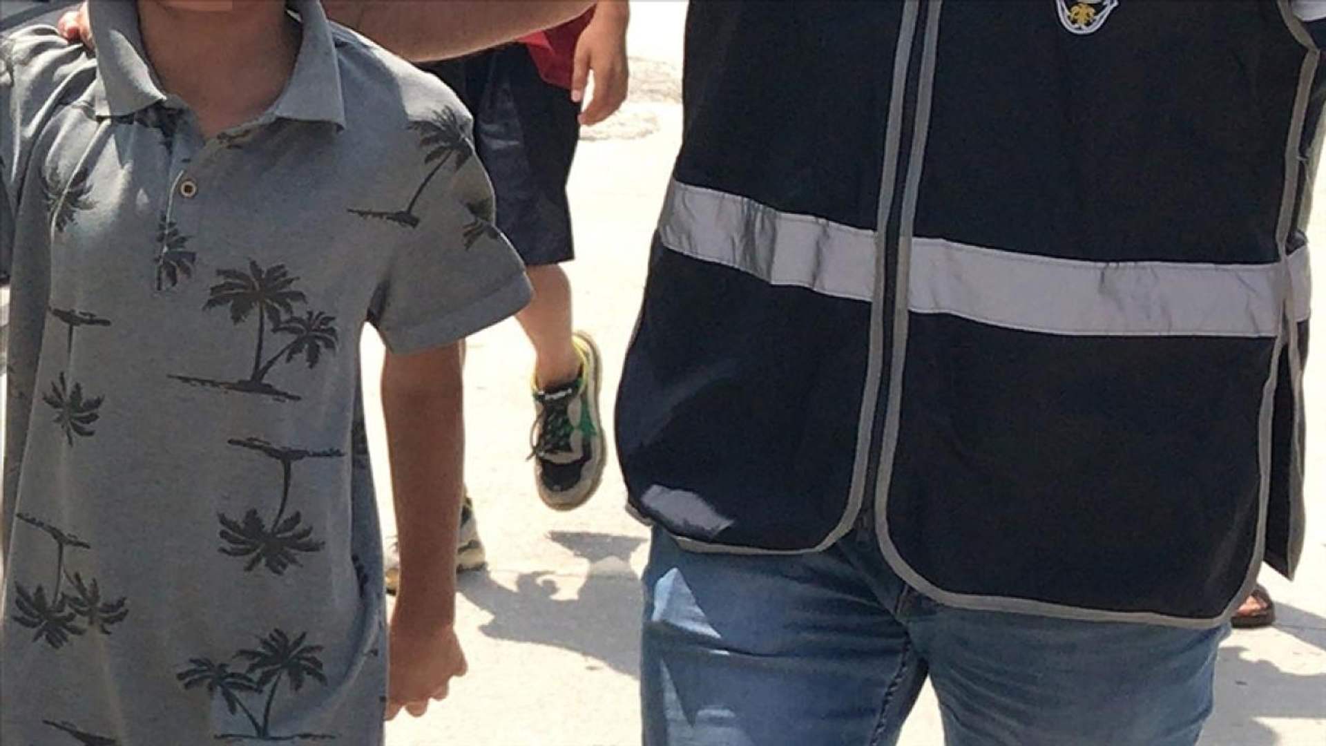 Yüzü görünmeyen bir çocuk, lacivert yelekli bir yetişkinin yanında güneşli bir yolda yürürken görülüyor.