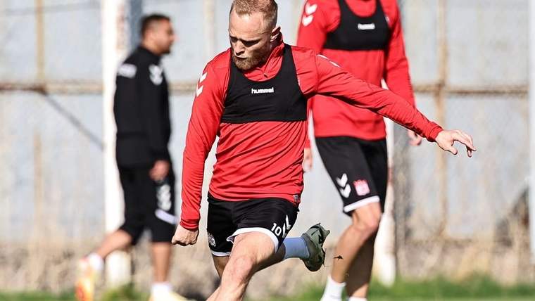 Kırmızı formalı bir futbolcu antrenman sahasında topa vururken, arka planda iki kişi bekliyor.