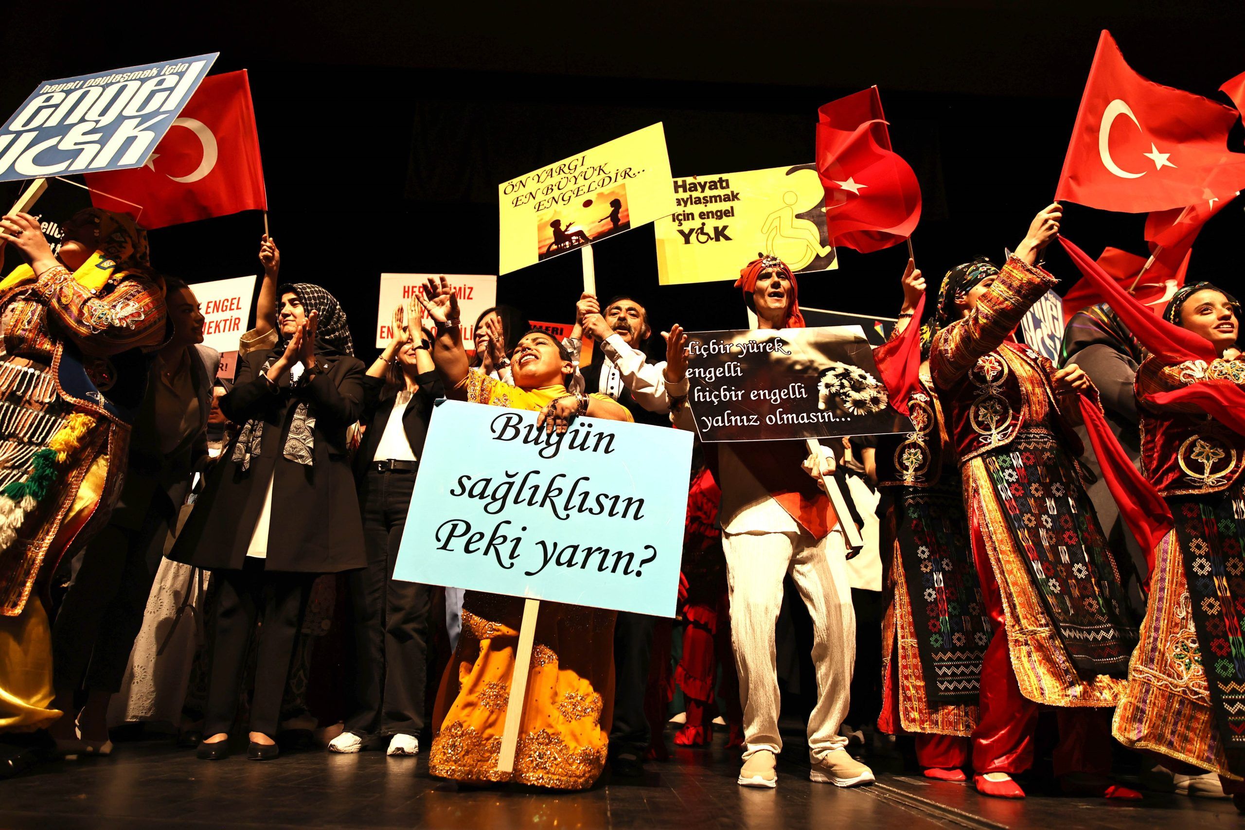 Sahne üzerinde çeşitli kostümler giymiş engelli bireyler, pankartlar tutarak gösteri yapıyorlar.