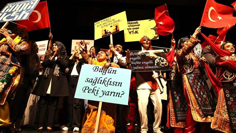 Sahne üzerinde çeşitli kostümler giymiş engelli bireyler, pankartlar tutarak gösteri yapıyorlar.
