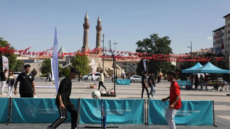 Açık alanda iki kişi, bariyerlerle çevrili sahada futbol topuyla karşılıklı dururken arkada cami minareleri görülüyor.
