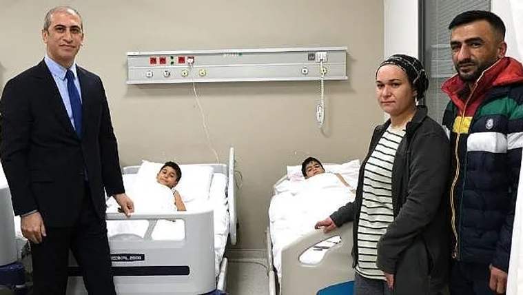 Hastane odasında iki çocuk yataklarda uzanırken, yanlarında üç yetişkin ayakta duruyor.