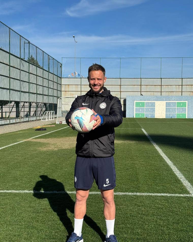 Futbol sahasında forma ve şort giyen bir kişi, ellerinde futbol topuyla kameraya bakıyor.